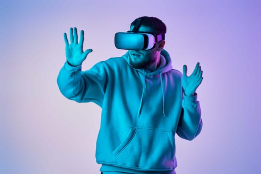 man in sweatshirt using virtual reality glasses and playing video games on blue background. Neon lighting