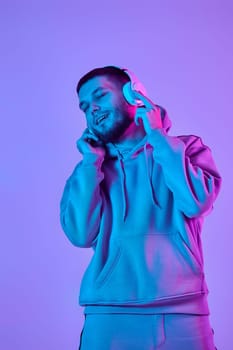 handsome bearded man with headphones in sweatshirt enjoying favorite music on purple neon background. Neon lighting