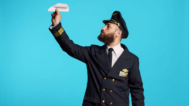 Young man working as pilot building paper plane, having fun with origami in studio. Male captain in aviation uniform playing with folded airplane, having professional occupation as airliner.