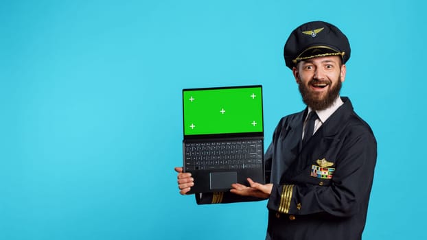 Happy young pilot holding laptop with greenscreen, using chroma key isolated display on portable computer. Aircrew captain in uniform looking at blank mockup template and copyspace.