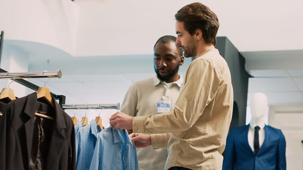 Shopping center worker helping client with trendy outfits to buy modern clothes in clothing store. Young shopper talking to employee and examining formal wear on hangers. Handheld shot.