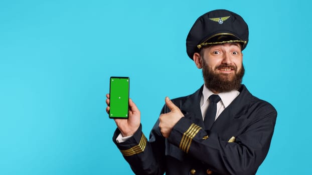 Smiling male aviator using telephone with greenscreen, holding smartphone with chroma key display and isolated template. Young airplane captain in flying uniform looking at mockup copyspace.