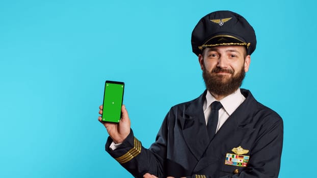 Young aviator using greenscreen dressed as captain, showing smartphone with isolated mockup display in studio. Professional flying pilot holding phone with blank chroma key template.