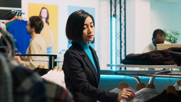 Young client shopping in fashionable boutique shop, looking at formal wear on hangers and racks. Woman buying trendy store merchandise from shopping mall, commercial activity.