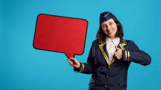 Positive woman holding red speech bubble on camera, working on commercial flights ad in studio. Smiling air hostess showing blank copyspace on carton board, flying industry job.