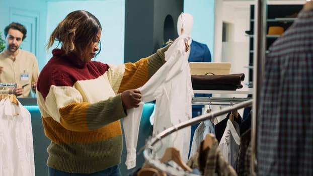 Retail store customer examining clothes on racks, looking to buy new trendy collection to increase wardrobe. Young woman checking clothing store merchandise in modern shopping mall.