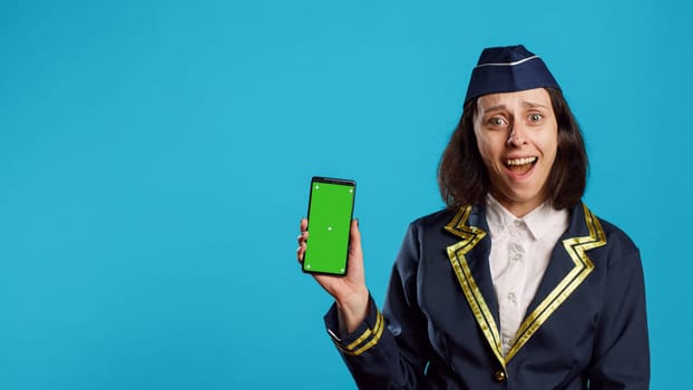 Positive aviation worker showing greenscreen on display, working with isolated mockup template on smartphone. Young happy stewardess using mobile phone with blank chroma key screen.