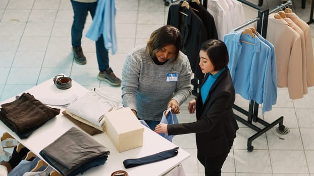 Group of diverse people examining trendy clothes in shop, checking discount on fashion collection. Retail store customers buying fromal or casual wear, commercial activity. Handheld shot.