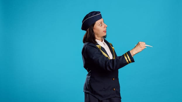 Caucasian woman playing with plane toy, being dressed as stewardess and showing miniature fake airplane in studio. Person working as air hostess holding artificial mini aircraft over blue background.