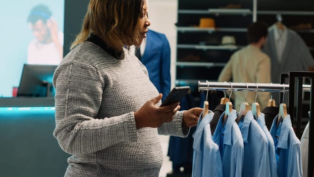 Female client checking online website stock in shop, trying to find formal wear from internet app in department store. Young adult using phone to buy favorite clothes in shopping center.