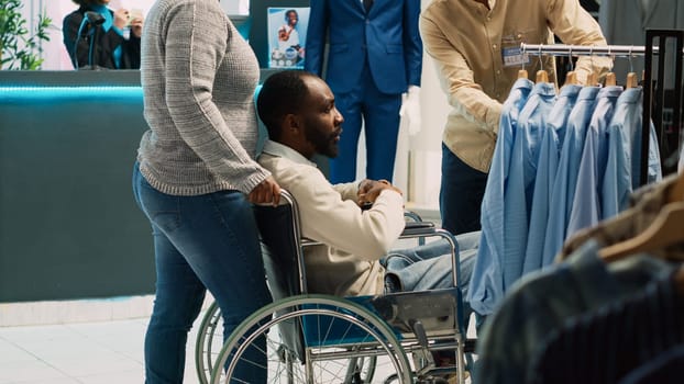 Young adult with impairment visiting clothing boutique, looking to buy modern casual wear in shopping mall. Male client wheelchair user examining fashionable clothes. Tripod shot.