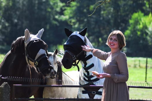 confident blonde woman feeding a horse among tall trees in summer,the countryside, rural, rustic scene, High quality photo