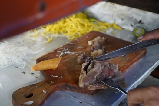 Beef brisket being chopped into bits on a wooden cutting board. with yellow noodle on the background. Preparation for beef noodle soup in Indonesia.