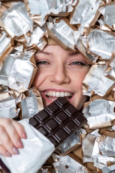 The face of a Caucasian woman surrounded by candy wrappers. The girl eats a bar of chocolate