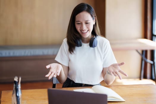 Portrait of a teenage Asian woman using a computer and notebook to study online via video conferencing on a wooden desk in library.