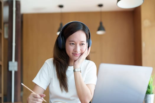 Portrait of a teenage Asian woman using a computer, wearing headphones and using a notebook to study online via video conferencing on a wooden desk in library.