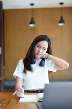 A portrait of a young Asian woman using a computer, wearing headphones and using a notebook to study online shows boredom and pain from video conferencing on a wooden desk in library.