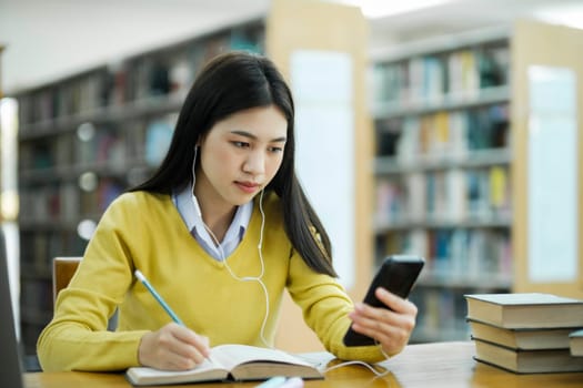 Young female college highschool student with headphones and in casual cloths sitting at desk studying, reading books, and writing with handphone at library for research or school project. E-Learning Education Library concept.