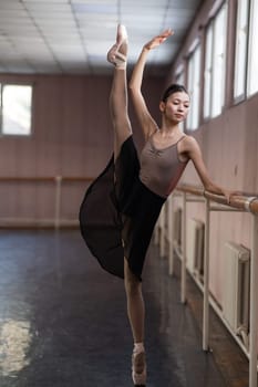 Graceful Asian ballerina in a beige bodysuit and black skirt is rehearsing in a dance class