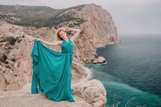 Side view a Young beautiful sensual woman in a mint long dress posing on a volcanic rock high above the sea during sunset. Girl on the nature on overcast sky background. Fashion photo