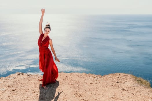 Side view a Young beautiful sensual woman in a red long dress posing on a rock high above the sea during sunrise. Girl on the nature on blue sky background. Fashion photo.