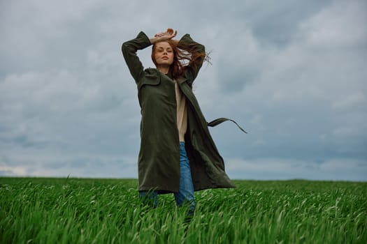 a woman in a long coat stands in a field against a cloudy sky with her back to the wind with her hands raised behind her head. High quality photo