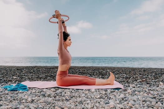 Middle aged well looking woman with black hair doing Pilates with the ring on the yoga mat near the sea on the pebble beach. Female fitness yoga concept. Healthy lifestyle, harmony and meditation.