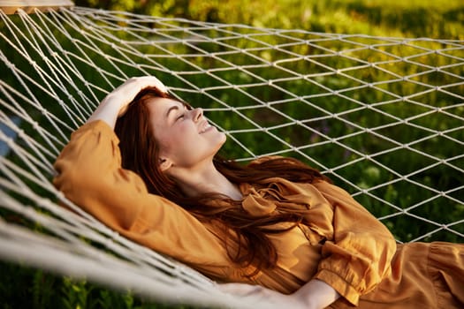 a happy woman is resting in a mesh hammock with her head resting on her hand, smiling happily looking away, enjoying a warm day in the rays of the setting sun, lying in an orange dress. High quality photo