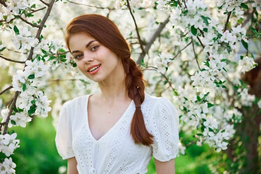beautiful red-haired woman in a white dress smiling stands near a flowering tree. High quality photo