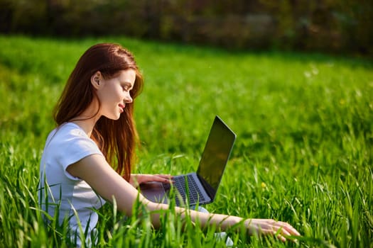 red-haired woman sits sideways to the camera in a field with a laptop in her hands. High quality photo