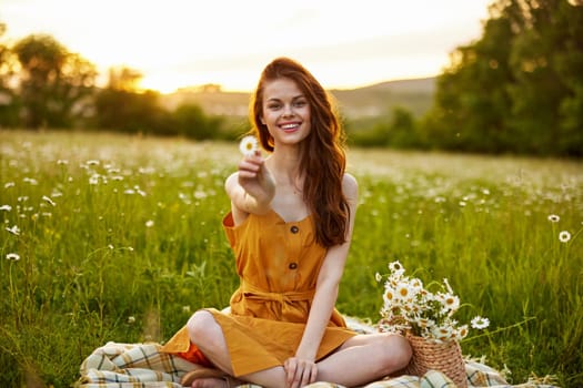 happy redhead woman sitting in a chamomile field on a plaid in a lotus position and holding a flower in her hands smiling at the camera. High quality photo