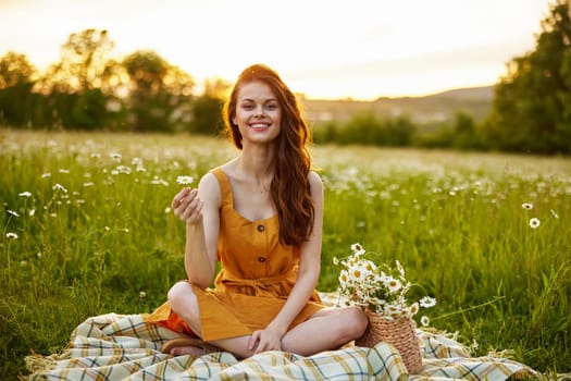 happy redhead woman sitting in a chamomile field on a plaid in a lotus position and holding a flower in her hands smiling at the camera. High quality photo