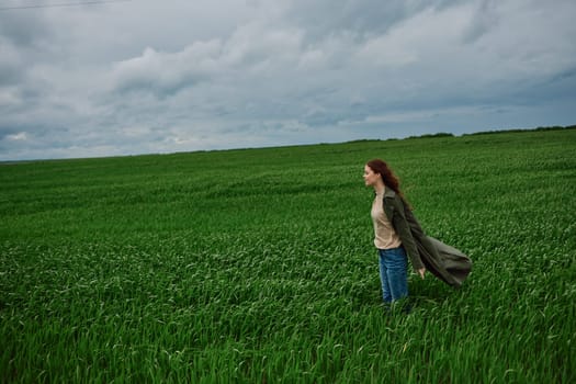 a beautiful woman stands in a green field in a raincoat and looks into the distance. Strong wind, flying hair, harmony with nature. High quality photo