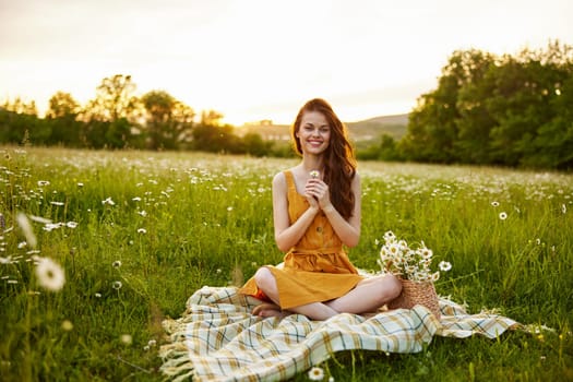 happy redhead woman sitting in a chamomile field on a plaid in a lotus position and holding a flower in her hands smiling at the camera. High quality photo