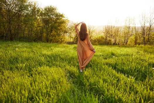 horizontal photo of a woman in an orange dress walking across the field holding her hand near her head, lifting it up, during the setting sun. High quality photo