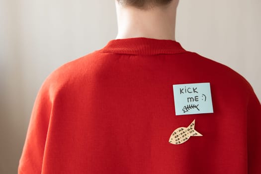 Boy in red hoodie with a paper attached with tape to his back with the handwriting text kick me. Fools day joke.