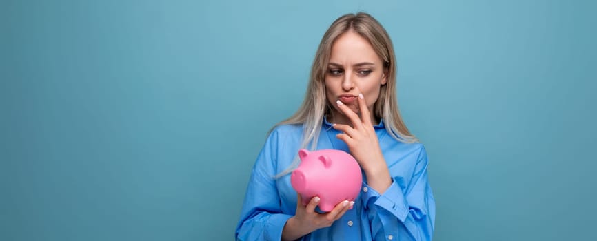 doubting blonde girl holding a piggy bank with pocket money and thinking where to spend the money on a blue isolated background.
