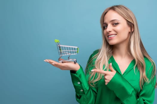 horizontal photo of stylish european woman shopper in green shirt with supermarket trolley on blue studio background with free space.