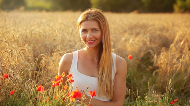 Young woman sitting in wheat field, lit by afternoon sun, few red poppies around her in foreground.