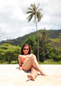 Young woman in bikini and sunglasses, sitting on the beach sand, smiling, palms behind her.