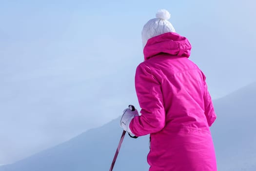 Look from behind, young woman in pink ski jacket, poles in her hands, getting ready to go down the hill.