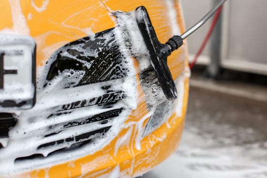 Yellow car front washed in self serve carwash, white shampoo foam contrasting with black front mask and brush.