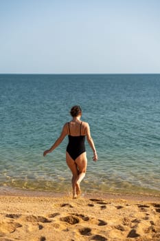 Woman sea swimsuit sand. A girl in full growth stands with her back and enters the sea in a black swimsuit. Alone on the beach on a sunny day