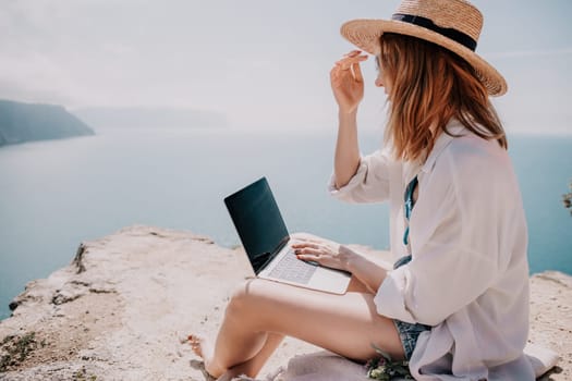 Successful business woman in yellow hat working on laptop by the sea. Pretty lady typing on computer at summer day outdoors. Freelance, travel and holidays concept.