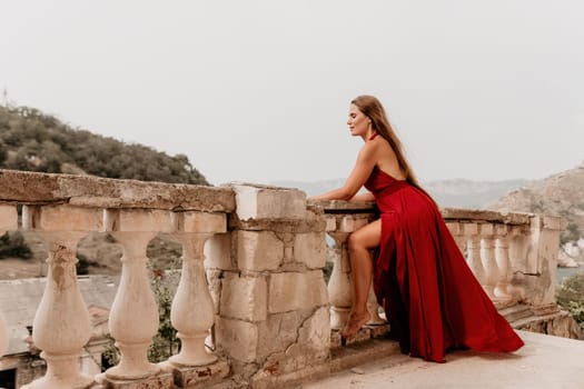 Side view a Young beautiful sensual woman in a red long dress posing on a volcanic rock high above the sea during sunset. Girl on the nature on overcast sky background. Fashion photo