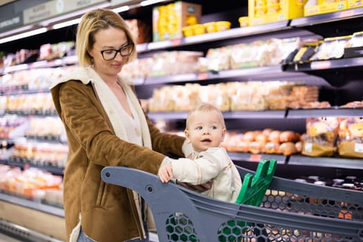 Caucasian mother shopping with her infant baby boy child choosing products in department of supermarket grocery store