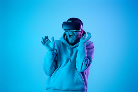 scared bearded man using virtual reality glasses and playing video games on blue background. Neon lighting