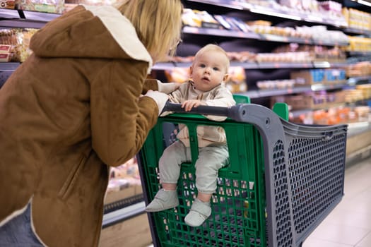 Caucasian mother shopping with her infant baby boy child choosing products in department of supermarket grocery store