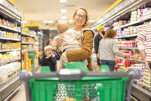 Mother shopping with her infant baby boy child, pushing shopping cart down department aisle in supermarket grocery store