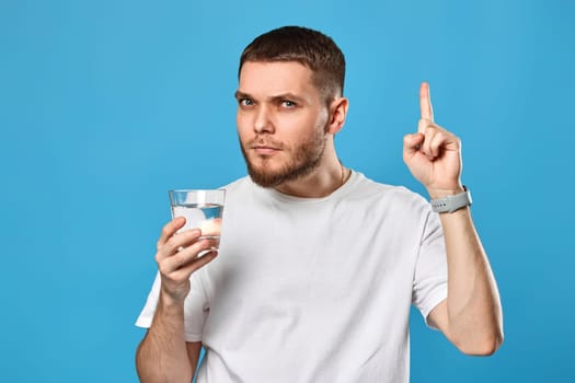 Young bearded man with drinking glass of water pointing up on blue background